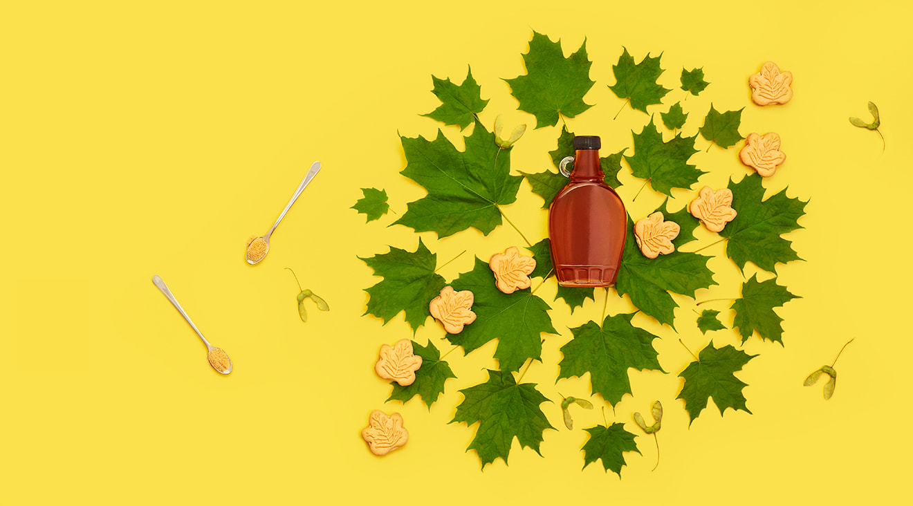 Maple syrup bottle surrounded by green maple leaves, maple cookies, and spoons filled with maple flakes.